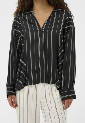 Vrouw draagt een zwart overhemd met witte verticale strepen en een witte broek met dunne zwarte verticale strepen.