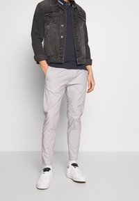 Muž v černé džískové bundě, námořnickém polo tričku, světle šedých kalhotách, bílých ponožkách a bílých teniskách stojící s rukama v kapsách.