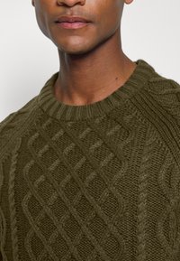 Suéter de punto verde oliva con cuello redondo, que presenta un patrón de trenzas texturizadas y acentos de canalé en el cuello y los hombros.