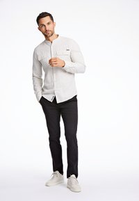 Hellgraues Button-down-Hemd mit zwei Brusttaschen, kombiniert mit schwarzen Hosen und weißen Turnschuhen. Das Hemd hat eine dezente Textur und einen abgerundeten Saum.