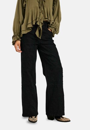 Persona que lleva pantalones negros de pierna ancha con bordado floral, una blusa verde oliva con volantes y zapatos de tacón beige con estampado.
