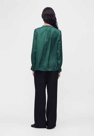 Femme aux longs cheveux foncés portant un chemisier vert brillant à manches longues et un pantalon noir à jambes larges, debout de dos sur un fond uni.