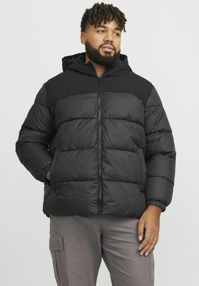 Geacă puffer neagră cu fermoar în față și glugă, având secțiuni quilted și manșete elastice, purtată peste pantaloni gri deschis.