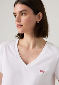 T-shirt blanc en V, fabriqué en coton, à manches courtes, avec un petit logo rouge Levi's sur la poitrine. Texture douce et coupe classique.