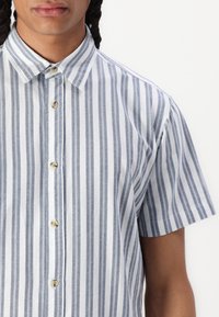Chemise à manches courtes à boutons avec des rayures verticales bleues et blanches, présentant un col pointu et des boutons beiges en matériau lisse.