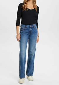 Frau, die ein schwarzes Langarmoberteil, hochgeschnittene blaue Jeans mit einem Vorderknopf und weiße Plateausneaker trägt, steht vor einem weißen Hintergrund.