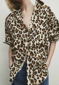 Chemise à imprimé léopard beige, ornée de taches marron et noires, dotée d'un col, de manches courtes et d'une coupe ample avec des boutons sur le devant et une poche poitrine.