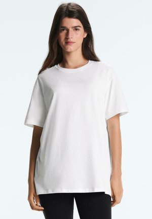 Junge Frau mit langen braunen Haaren, die ein schlichtes, weißes, übergroßes T-Shirt mit kurzen Ärmeln und schwarze Hosen trägt und vor einem einfarbigen Hintergrund steht.