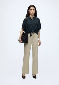 Femme aux longs cheveux foncés porte une chemise noire à manches courtes nouée, un pantalon beige taille haute, des talons noirs et porte un sac à main noir.