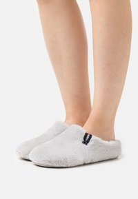 Zapatillas de casa grises y peludas con una textura suave, diseño de parte trasera abierta y acento de logo negro en el lateral. Adecuadas para uso en interiores.