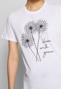 Camiseta blanca de algodón con un gráfico negro de tres flores parecidas a diente de león y el texto "florece con gracia" en una fuente manuscrita.