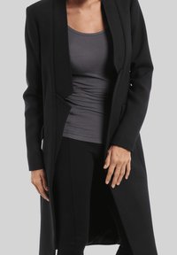 Abrigo largo negro con cuello chal, diseño sin costuras, bolsillos laterales y detalle texturizado. Lleva puesto sobre una camiseta gris ajustada.