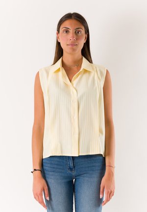 Giovane donna con capelli lunghi e scuri indossa una camicia senza maniche giallo pallido con bottoni e jeans blu, in piedi davanti a uno sfondo chiaro e uniforme.