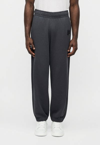 Άνδρας που φοράει σκουρόχρωμο γκρι τζόγκερ με ελαστική μέση και μανσέτες στους αστραγάλους, καθώς και λευκά αθλητικά παπούτσια, στέκεται μπροστά σε απλό λευκό φόντο.