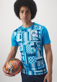 Tricou turcoaz sportiv cu modele geometrice, mâneci scurte, guler tip crew și un logo, ținut împreună cu o minge de fotbal portocalie și gri.
