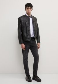 Giovane uomo che indossa una giacca di pelle nera, una camicia bianca, jeans scuri e scarpe nere, in piedi contro un muro chiaro e semplice.