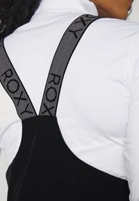 Schwarzes Kleidungsstück mit breiten, strukturierten Trägern, die mit fetter Schrift versehen sind, kombiniert mit einem figurbetonten weißen Hemd. Fokus auf sauberer Naht und glatten Oberflächen.