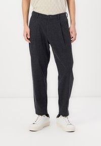 Pantaloni din lână gri cu pliere, picior îngustat și închidere cu nasture. Purtați cu adidași albi. Suprafața are o textură subtilă.