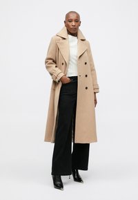 Persona in abito lungo doppiopetto beige, maglione bianco, pantaloni neri a gamba larga e stivali neri a punta, in piedi su uno sfondo neutro.