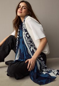 Eșarfă cu un model floral în nuanțe de albastru și alb, fabricată dintr-un material texturat, purtată de un model, asociată cu un top alb și blugi negri.