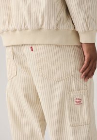 Μπεζ ριγέ παντελόνια από βαμβάκι, με τσέπη με πατς και κόκκινο λογότυπο, ενισχυμένες ραφές και ραβδωτή μέση.