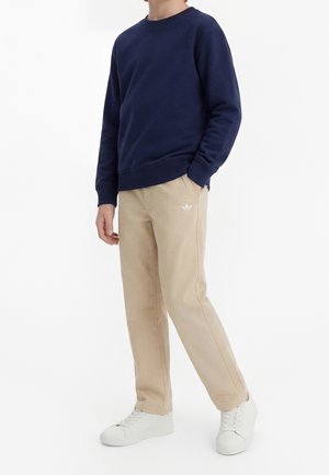Persona con sudadera azul marino, pantalones beige de Adidas con logo en el bolsillo y zapatillas blancas, de pie con una mano en el bolsillo.