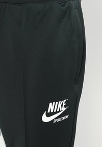 Svarta idrottsbyxor i mjukt tyg, med en vit Nike-logotyp och texten "SPORTSWEAR", sidofickor och elastisk midja.