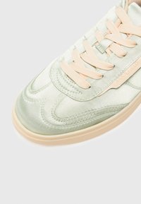 Baskets satin vert clair avec des lacets beiges, présentant un revêtement texturé et des détails de couture subtils. Semelle en caoutchouc de couleur nude.