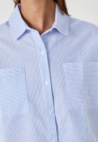 Camicia a righe blu e bianche con colletto aperto, dotata di due tasche frontali con righe orizzontali e un tessuto testurizzato.
