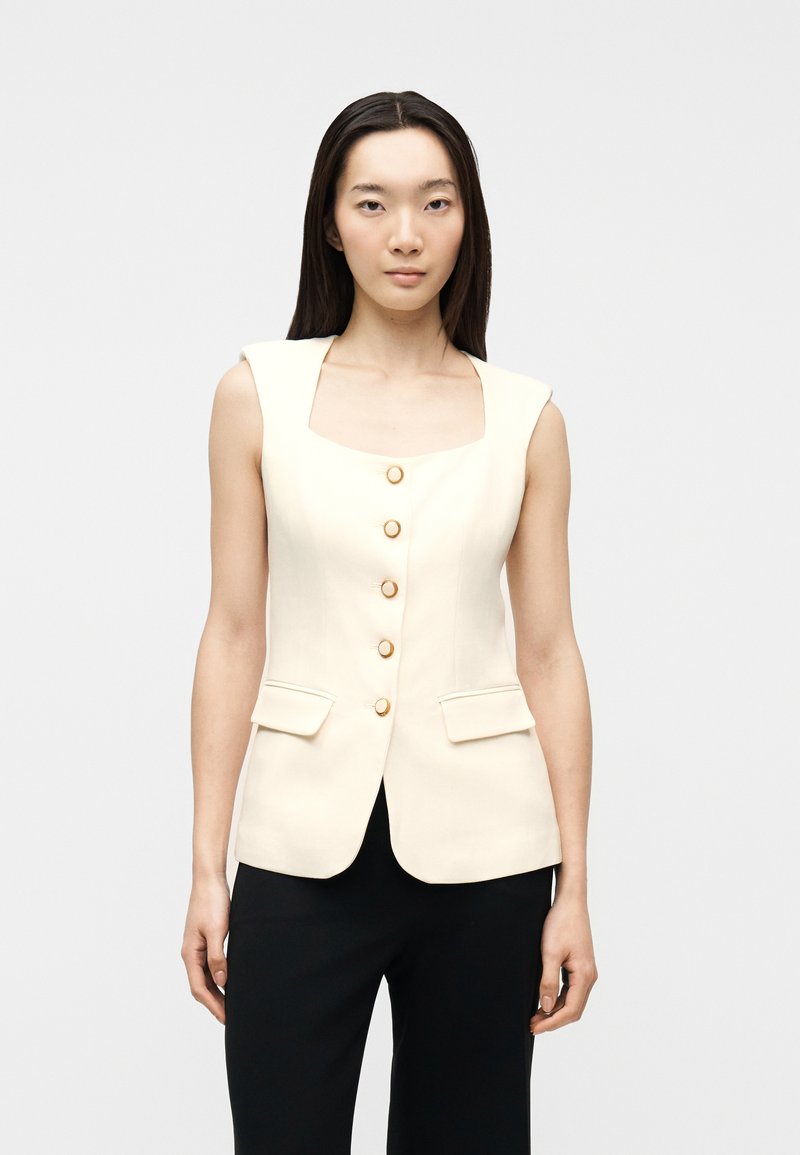 Γυναίκα που φοράει αμάνικο εκρού γιλέκο με χρυσά κουμπιά και τσέπες με καπάκι, συνδυασμένο με μαύρο παντελόνι, στέκεται μπροστά σε απλό φόντο.