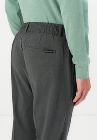 Grå bukser med elastisk midje og en glatt tekstur, med en baklomme og et merke. Komplementert av en lys grønn genser.