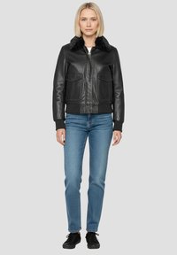 Giacca bomber in pelle nera con collo di pelliccia, tasche frontali con patta, polsini e orlo a coste, abbinata a jeans azzurri e sneakers nere.