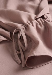 Materialul de culoare mov deschis prezintă un design plisat cu legături la mânecă, evidențiind o textură netedă și detalii minime de cusătură.
