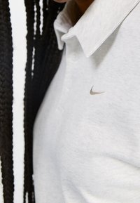 Ljusgrå Nike-polo med krage, tre knappar och en liten broderad logotyp i en något mörkare nyans. Strukturerad tygsynlighet.