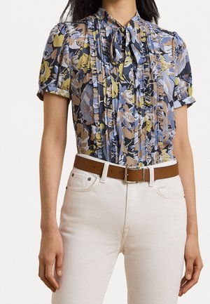 Mujer con blusa floral de manga corta con volantes y lazo, combinada con pantalones blancos y cinturón marrón.