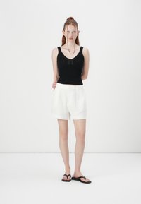 Top in maglia nera, pantaloni corti in lino bianchi e infradito nere. L'outfit ha un design semplice e casual con una palette di colori neutri.