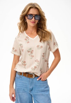 Kobieta z długimi blond włosami, nosząca duże kwadratowe okulary przeciwsłoneczne, bluzkę w paski w kwiaty, brązowy pasek oraz jasnoniebieskie dżinsy z jedną ręką w kieszeni.