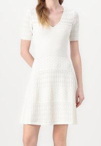 Robe blanche en tricot avec manches courtes, col en V, texture supérieure en dentelle et motifs de points en étages sur la jupe. Corsage ajusté avec ourlet évasé.