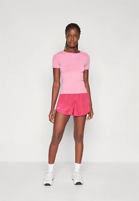 Mujer de pie contra un fondo blanco, vestida con una camiseta deportiva de manga corta rosa, pantalones cortos rojos, calcetines blancos y zapatillas blancas.