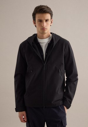 Joven con chaqueta negra con cremallera y capucha sobre una camisa blanca, de pie con una mano en el bolsillo contra un fondo liso.