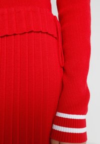 Rotes gestricktes Oberteil mit Rippenstruktur. Besitzt einen gewellten Saum und weiße Streifen an den Ärmelbündchen. Figurbetonter Schnitt.