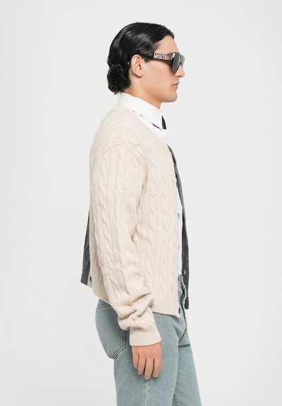Beige kabelstickad cardigan med en grå rygg, bärs över en vit skjorta och svart slips, i kombination med blå jeans och överdimensionerade svarta solglasögon.