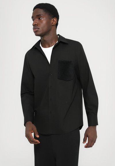 Homme portant une chemise noire à boutons avec une poche poitrine texturée et un t-shirt blanc en dessous, regardant légèrement sur le côté devant un fond uni.