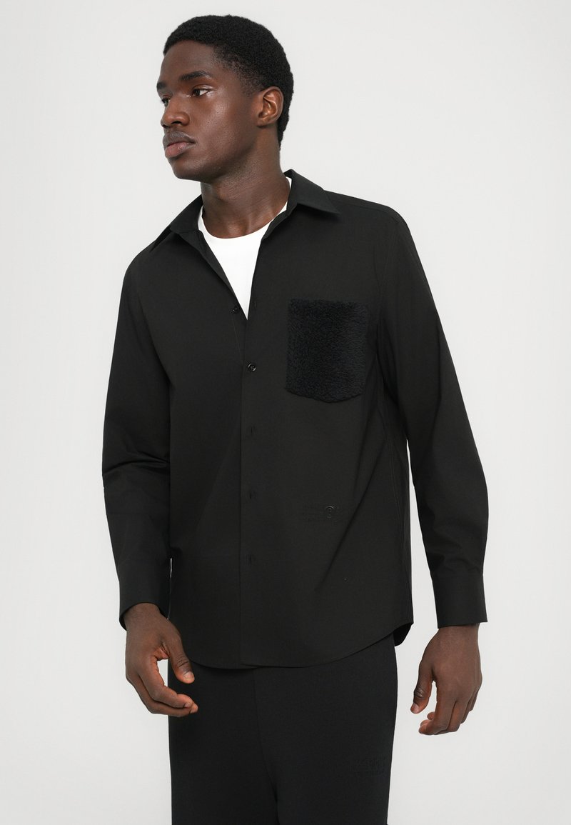 Mand iført en sort skjorte med knapper og tekstureret brystlomme samt en hvid undertrøje, der ser lidt til siden mod en ensfarvet baggrund.