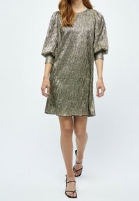 Metallisk grön-guld klänning med plisserad textur, rund halsringning och puffärmar, bärs med svarta sandaler med remmar.