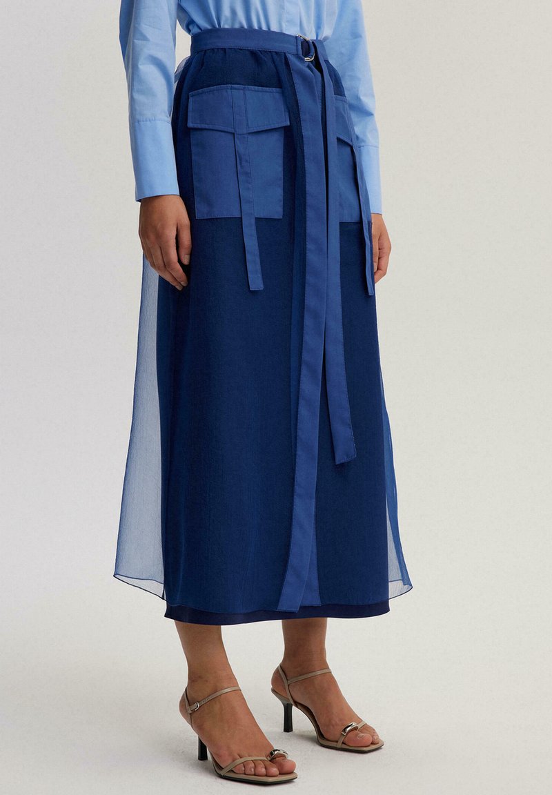 Sinine pikk seelik poolläbipaistva pealiskihiga, kahe esitasku ja sobiva lipsvööga. Omadusteks on sile tekstuur ja kihiline disain.