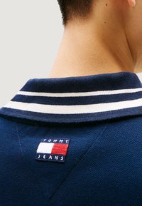 Polo blu navy con colletto a righe e patch con il logo Tommy Jeans nei colori rosso, bianco e blu sul retro. Tessuto strutturato.