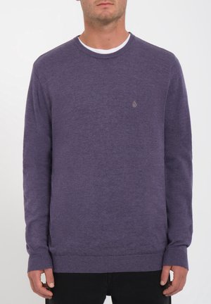 Homme portant un pull violet uni à col rond avec un petit logo sur le côté gauche de la poitrine, associé à un pantalon noir.