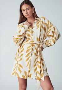 Abito camicia bianco con motivi a foglia gialla, maniche lunghe, bottoni sulla parte anteriore e una cintura coordinata in vita.