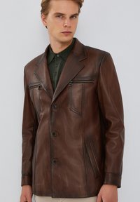Blazer de cuero marrón con cuello con muesca, dos bolsillos frontales y un bolsillo en el pecho con cremallera, con una textura suave y un ajuste entallado.